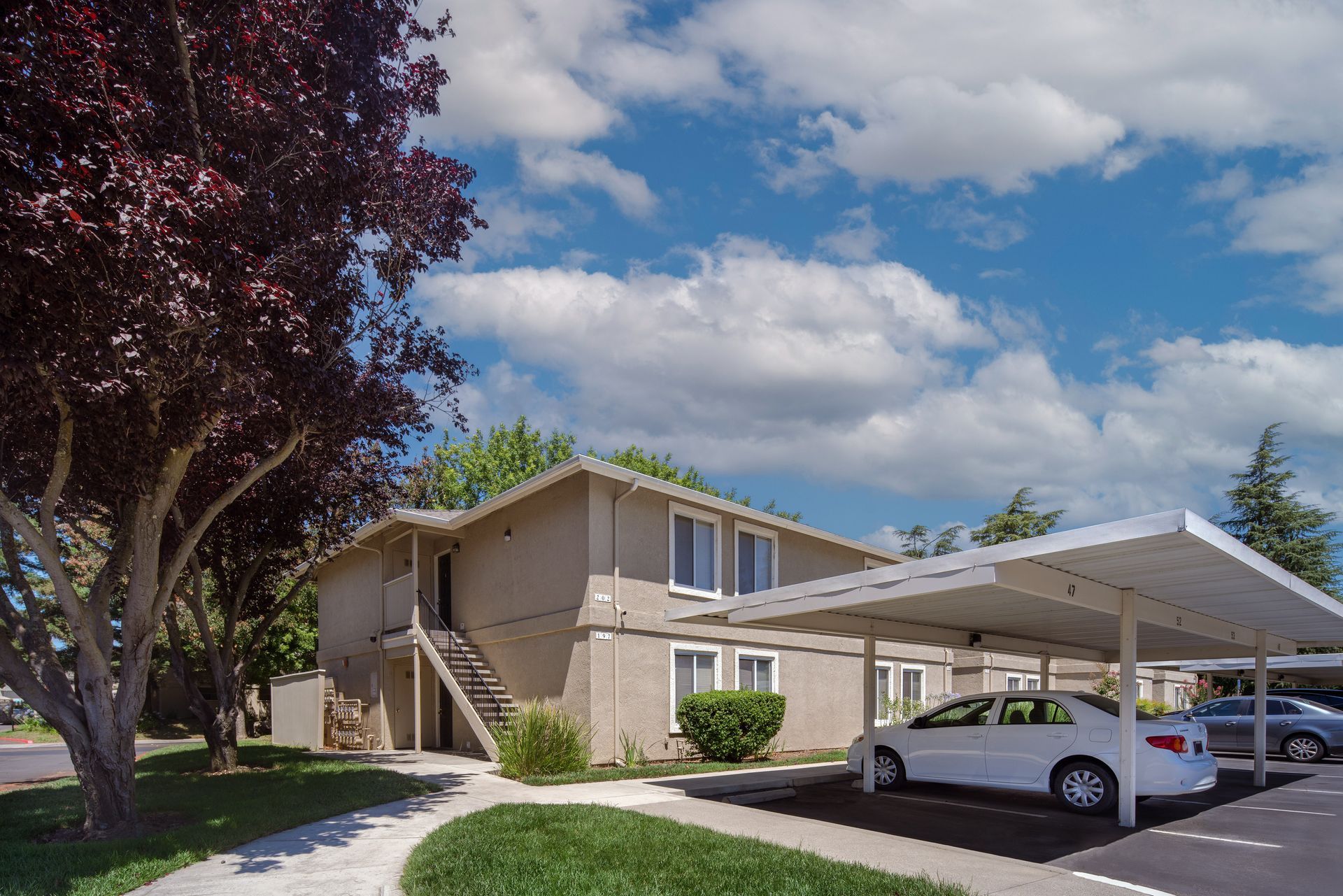 Apartment building with covered parking, cars, green lawn, and a cloudy blue sky.
