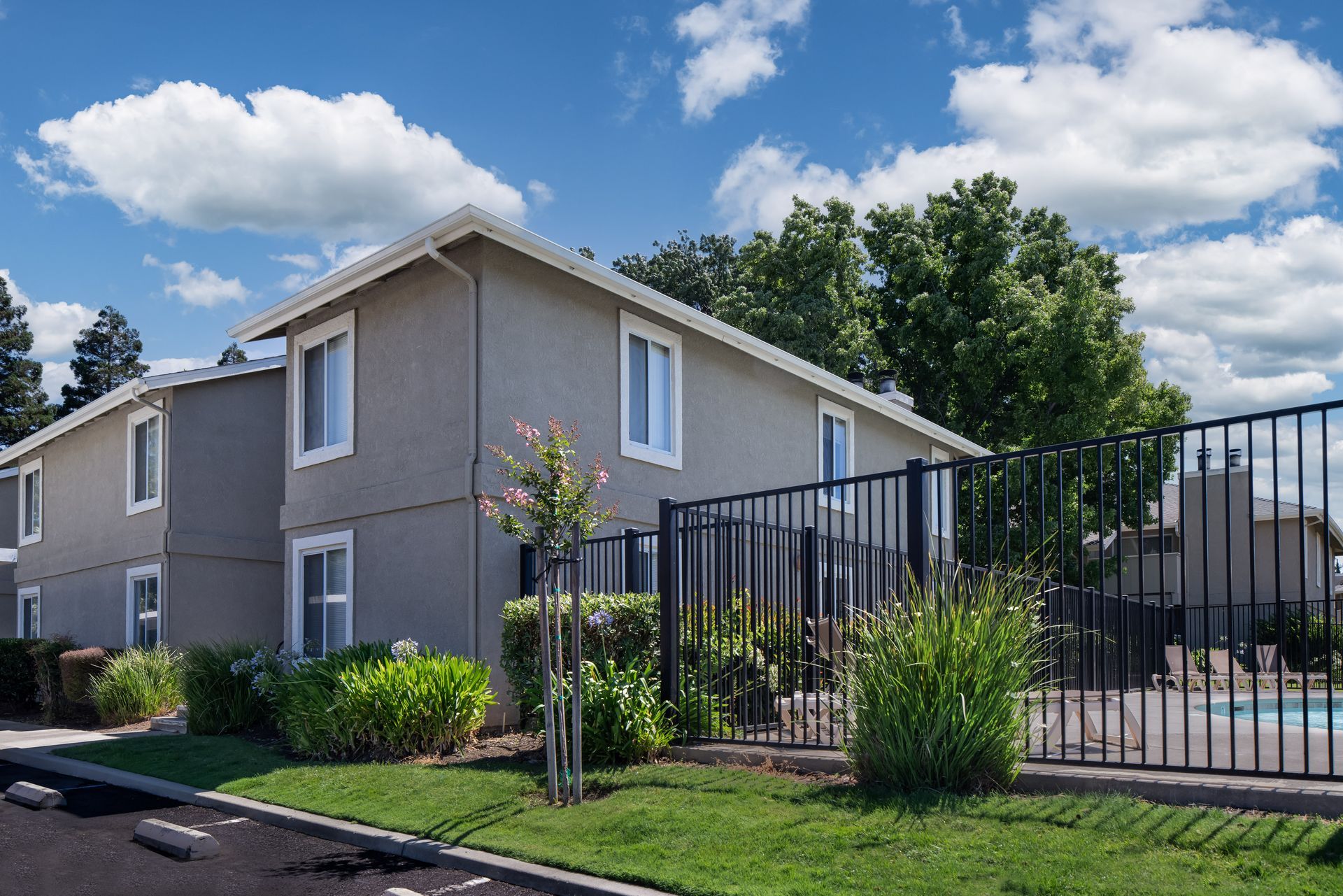 Two-story apartment buildings with a black fence, trees, and a pool under a blue sky.