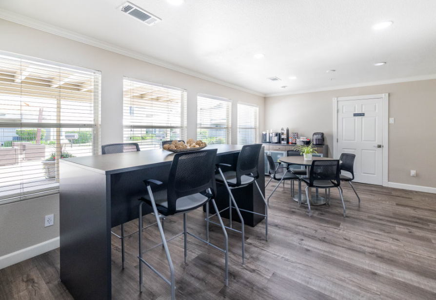 Community room with dark wood floors, windows, tables, chairs, and coffee station.