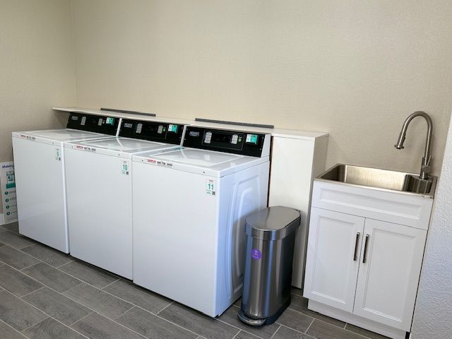 Laundry room with three white washing machines, a stainless steel sink, and a trash can.