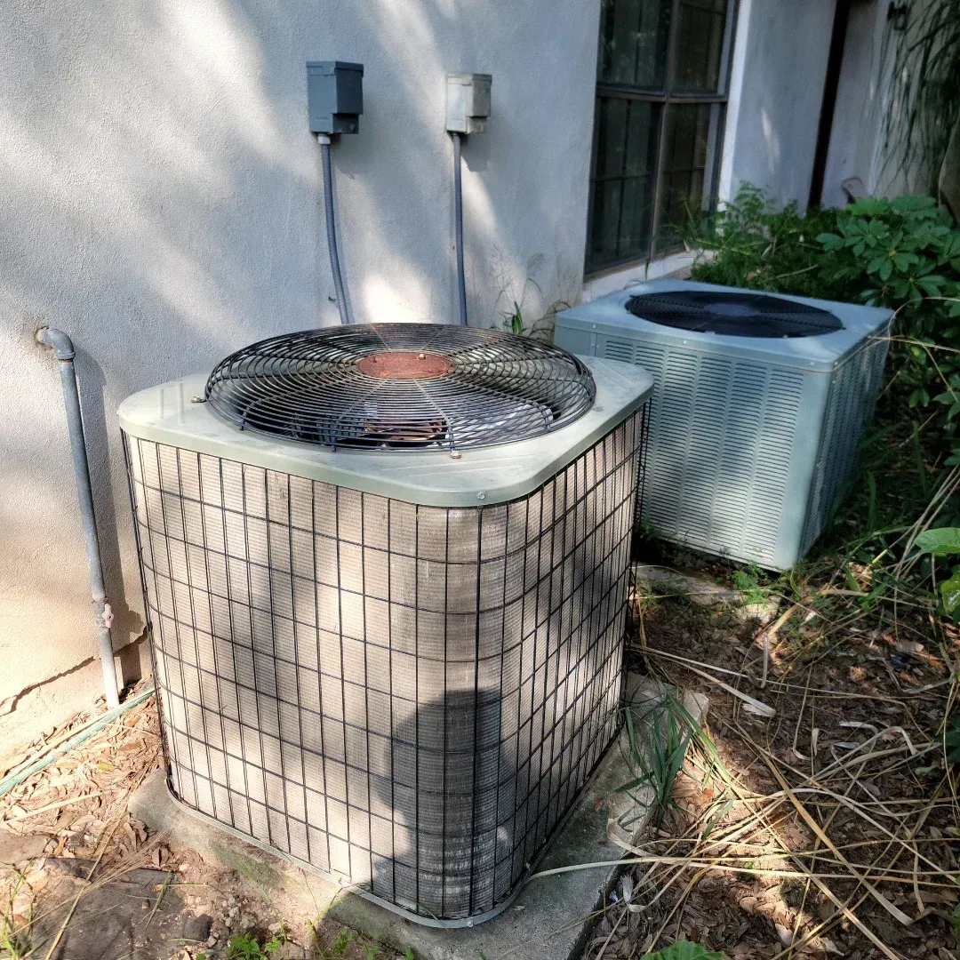 Two air conditioners are sitting outside of a house.