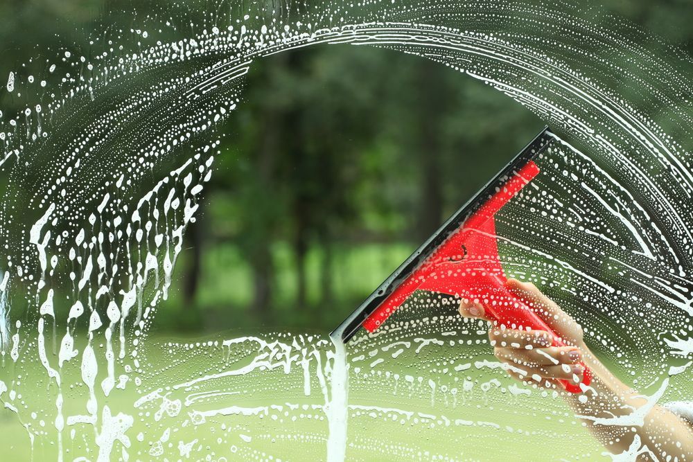 A person using a red squeegee to clean a soapy window; trees visible in the background — Totally Green Cleaning Solutions in Kambah, ACT