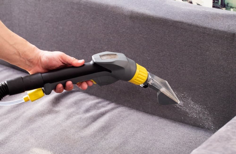 Person cleaning a gray couch with a professional upholstery cleaner, removing dust — Totally Green Cleaning Solutions in Kambah, ACT
