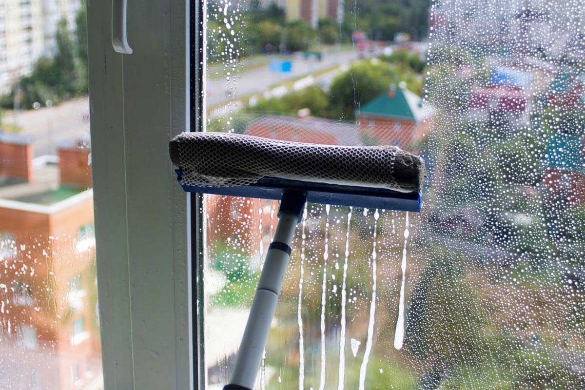 Window being cleaned with a squeegee, streaks of soapy water running down. View of buildings in the background — Totally Green Cleaning Solutions in Kambah, ACT