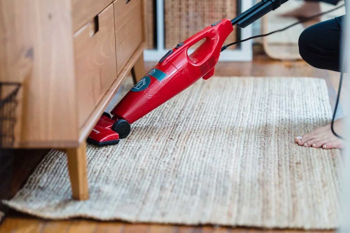 Red Vacuum Cleaner Cleaning a Rug Under a Wooden Dresser — Totally Green Cleaning Solutions in Kambah, ACT