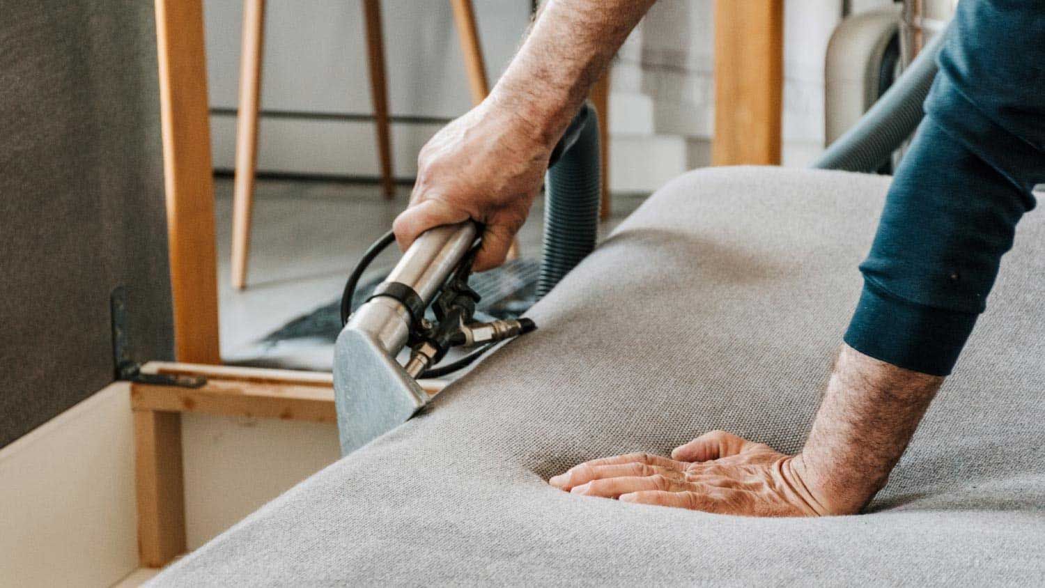 A Man Is Using a Steam Cleaner on A Grey Upholstered Couch — Totally Green Cleaning Solutions in Belconnen, ACT