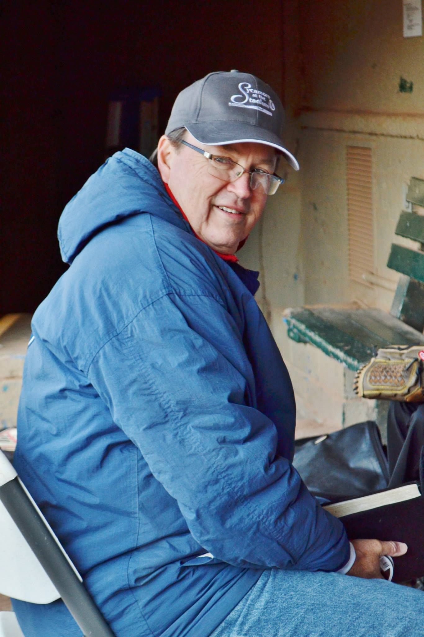 Man in blue jacket and cap, seated outdoors, smiling, holding a Bible.