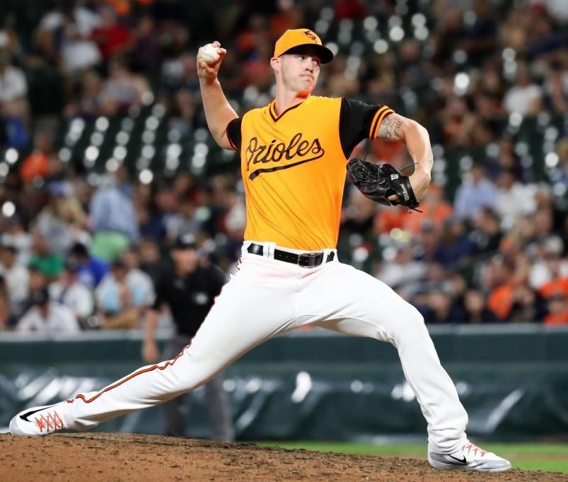 A baseball pitcher in orange jersey throws a ball; white pants; Orioles logo.