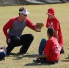A baseball coach kneeling, demonstrating glove catching to two young players in red shirts on a field.