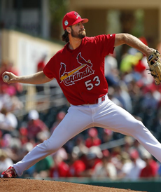 Baseball pitcher in red Cardinals uniform throwing a ball. White pants, red cap, sunny outdoor setting.
