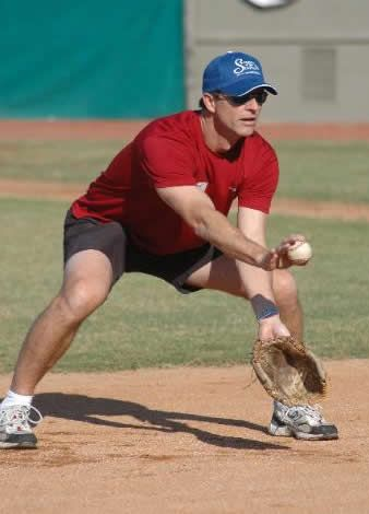 Man in red shirt and shorts fielding a baseball on a dirt field, wearing a blue cap and sunglasses.
