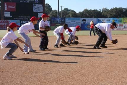 Children and adults practice fielding on a baseball field. Wearing red hats and white shirts, they are bent over, hands out.