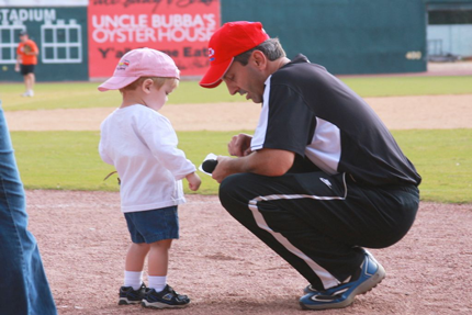Man in red cap kneeling, talking to a toddler on a baseball field.