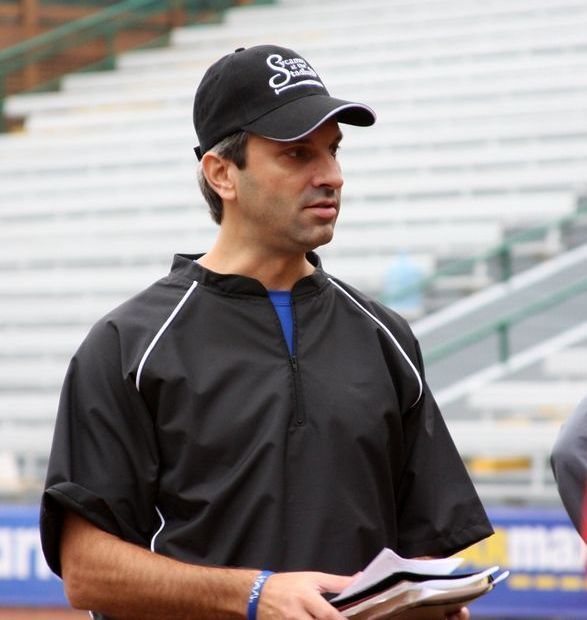 Man in black cap and jacket holding papers, looking to the side at a baseball field.