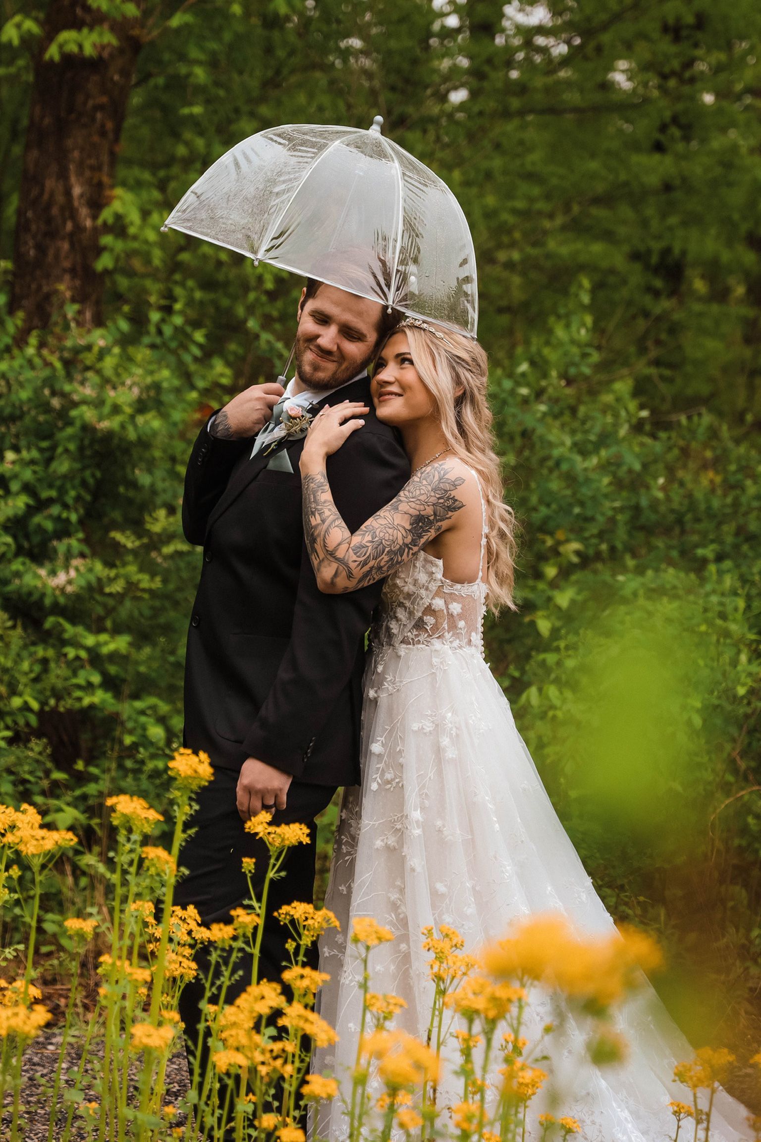 A couple in wedding attire stands under a clear umbrella amidst a forest of green trees and yellow wildflowers.