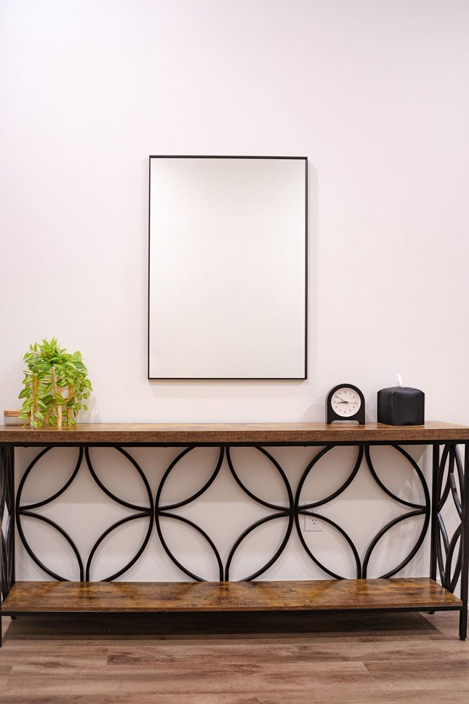 Minimalist console table with decorative metal front, mirror, small plant, and two clocks against a white wall.