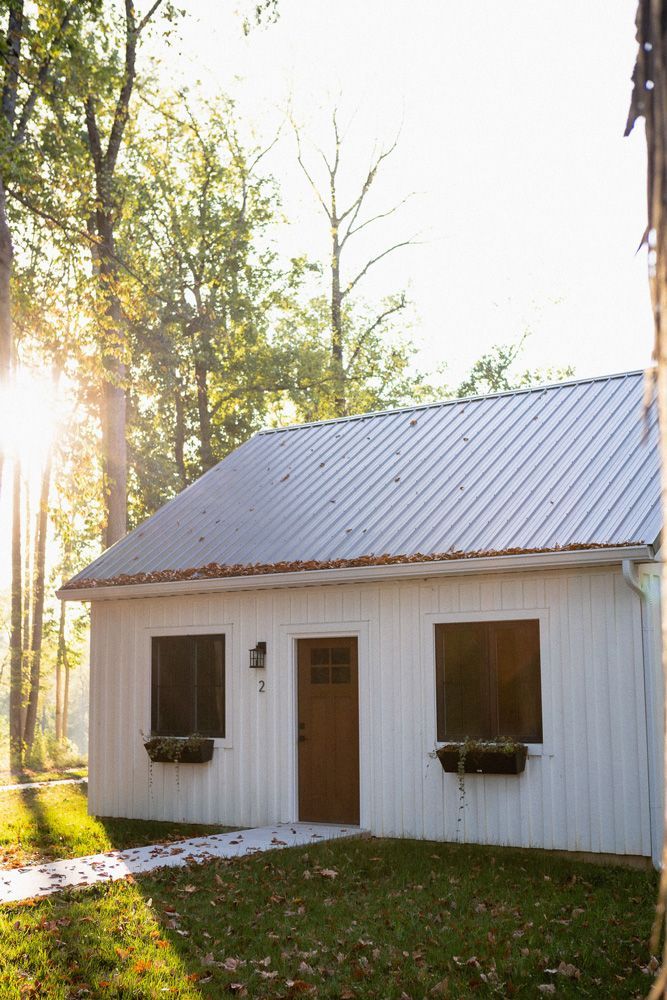 Small white cottage with a metal roof in a sunlit wooded yard