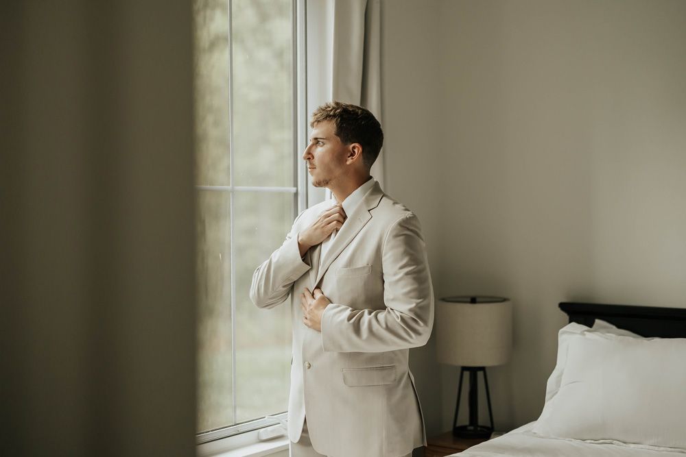 Man in a beige suit adjusting his collar beside a window in a softly lit bedroom
