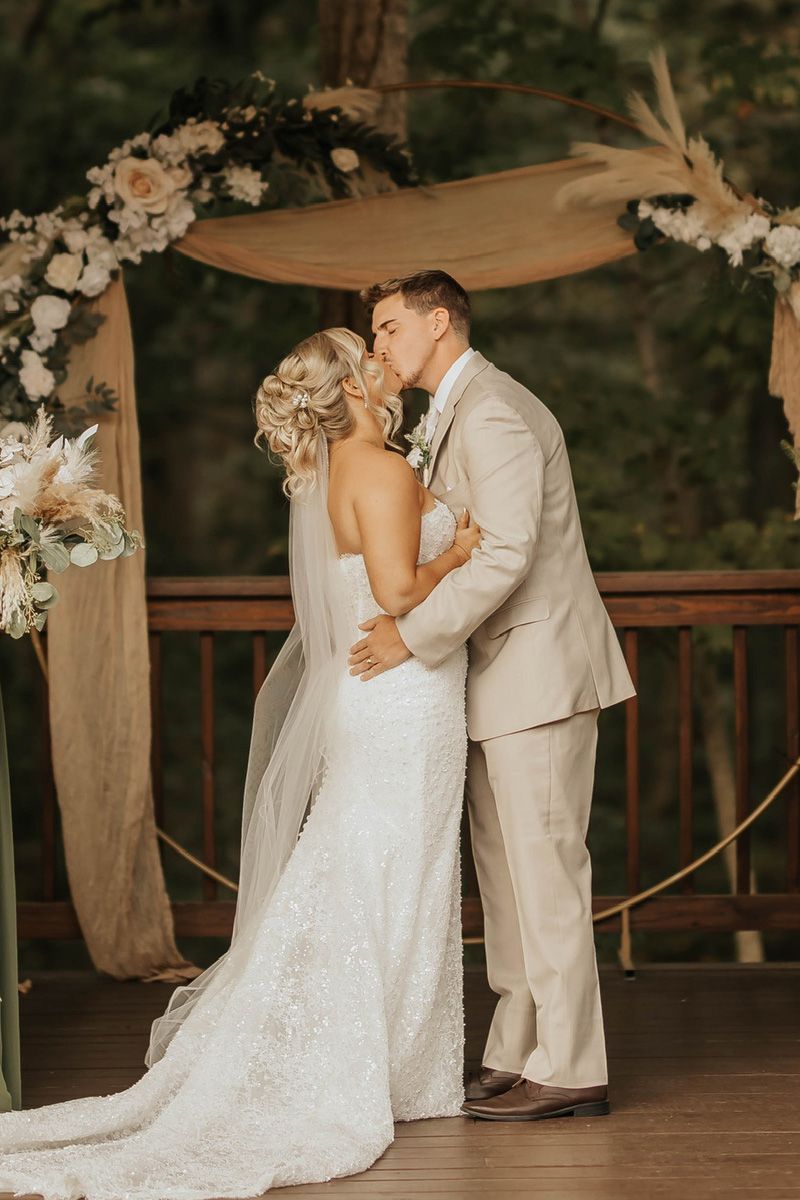 A bride and groom in wedding attire kiss under a floral arch on a wooden deck surrounded by trees.