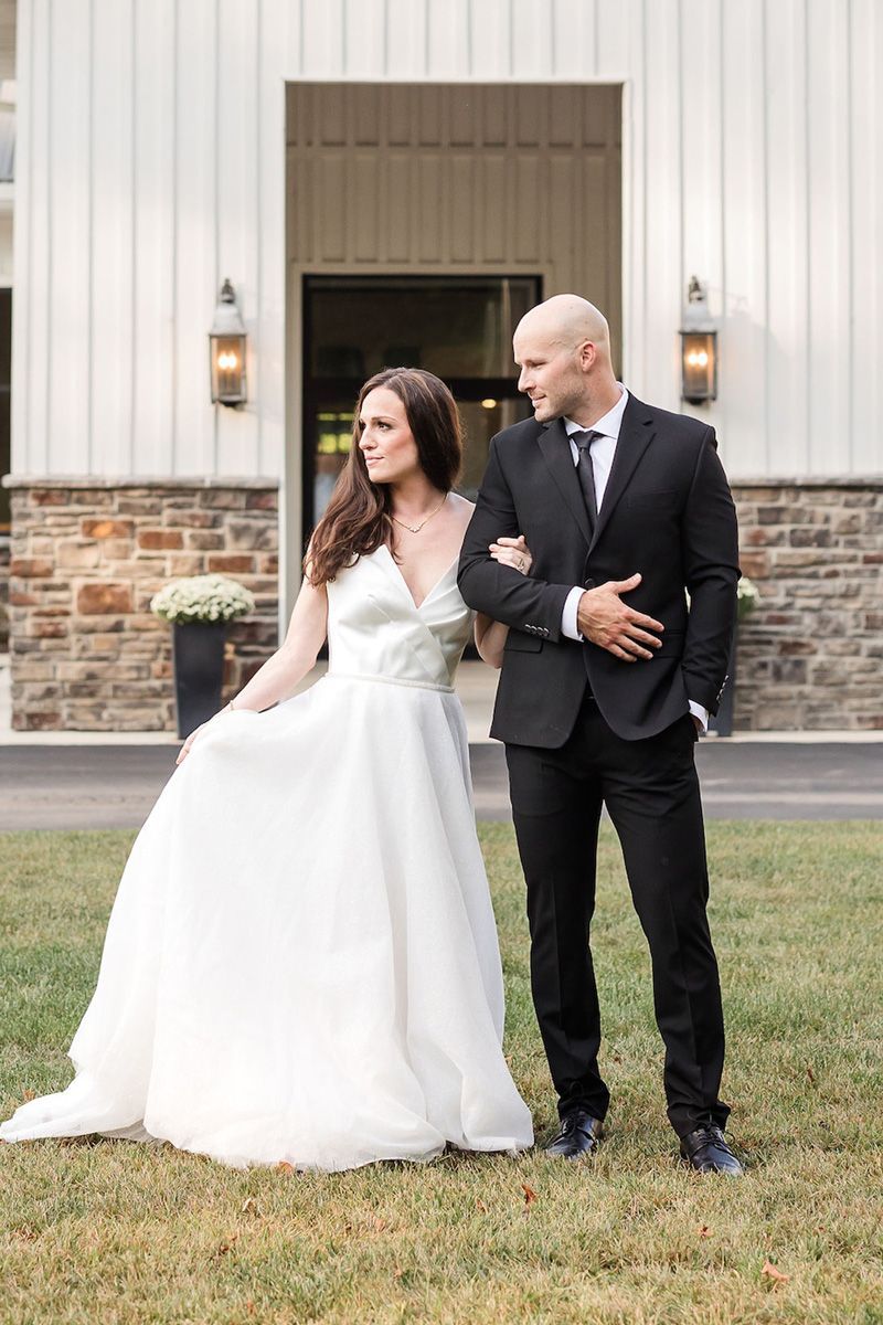 A bride in a white wedding gown holding her dress walks arm-in-arm with a person in a black suit outside a barn venue.