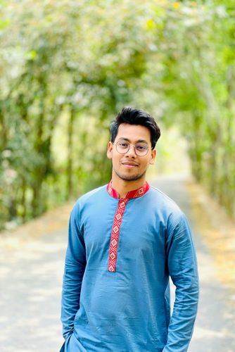 Person in a blue embroidered kurta standing on a tree-lined path, with a slight smile.