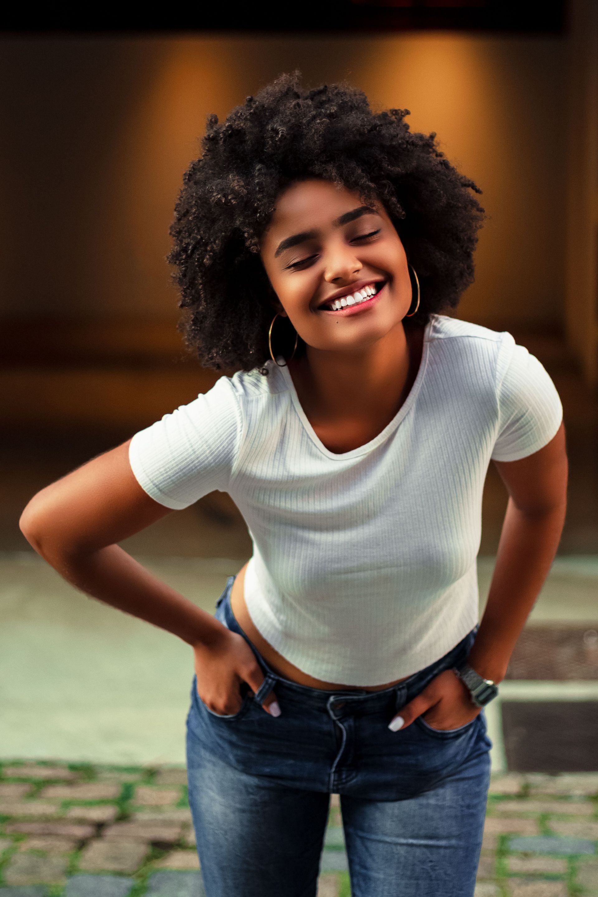 Smiling person in a white T-shirt and jeans leaning forward with hands on hips outdoors.