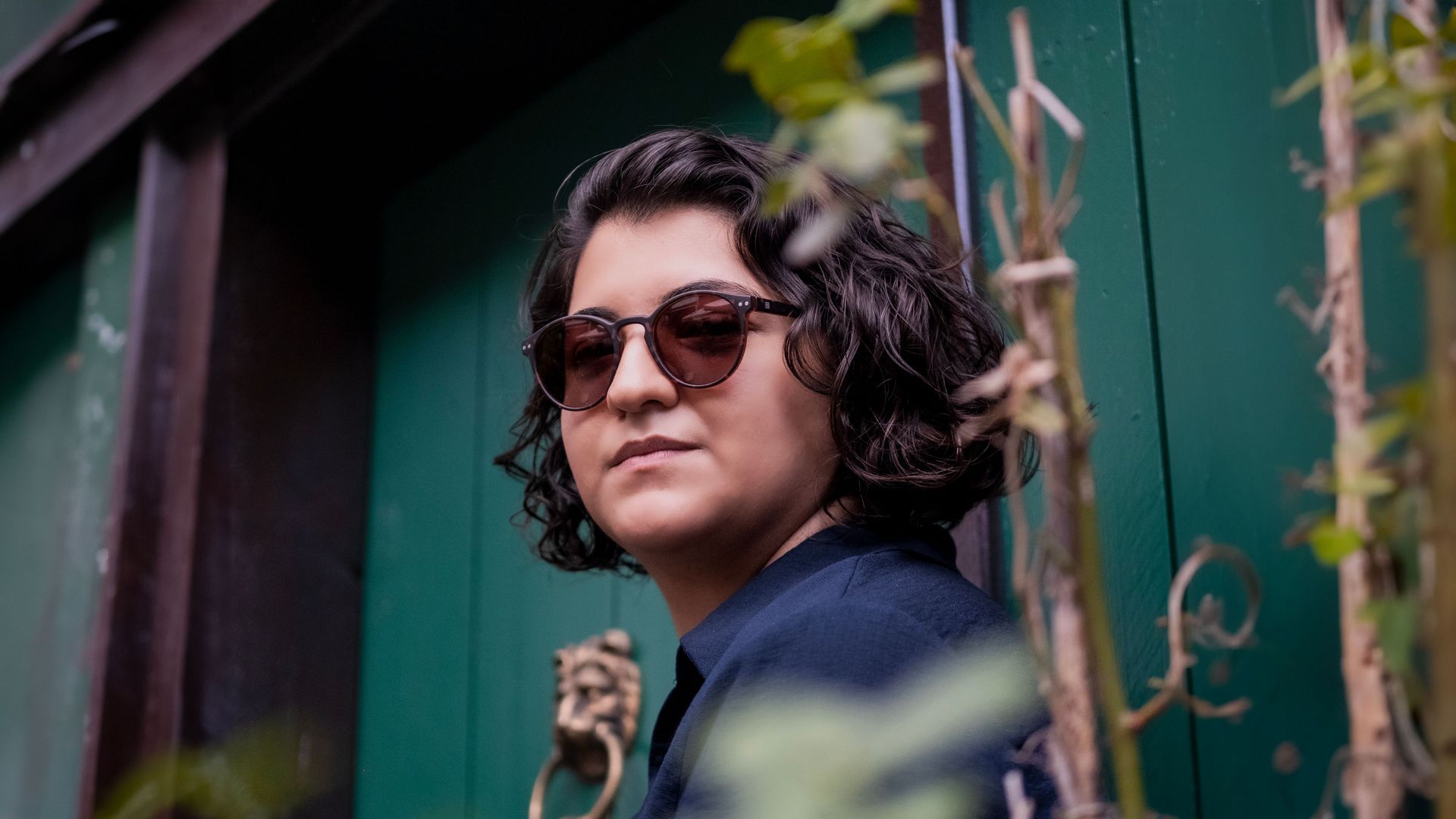 Person with curly dark hair and sunglasses stands by a green fence outdoors.