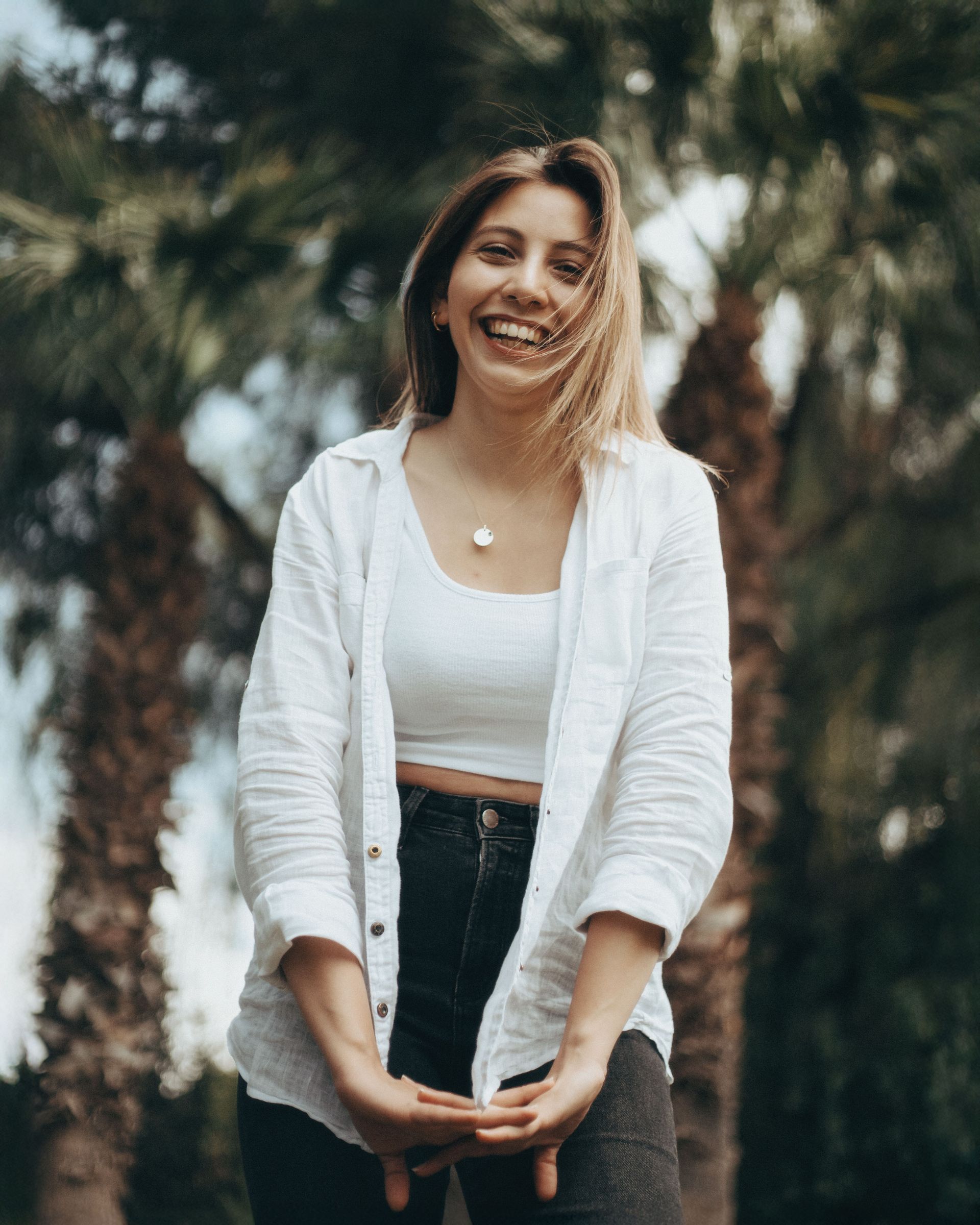 Smiling woman in a white cardigan and black jeans standing outdoors with blurred palm trees behind her