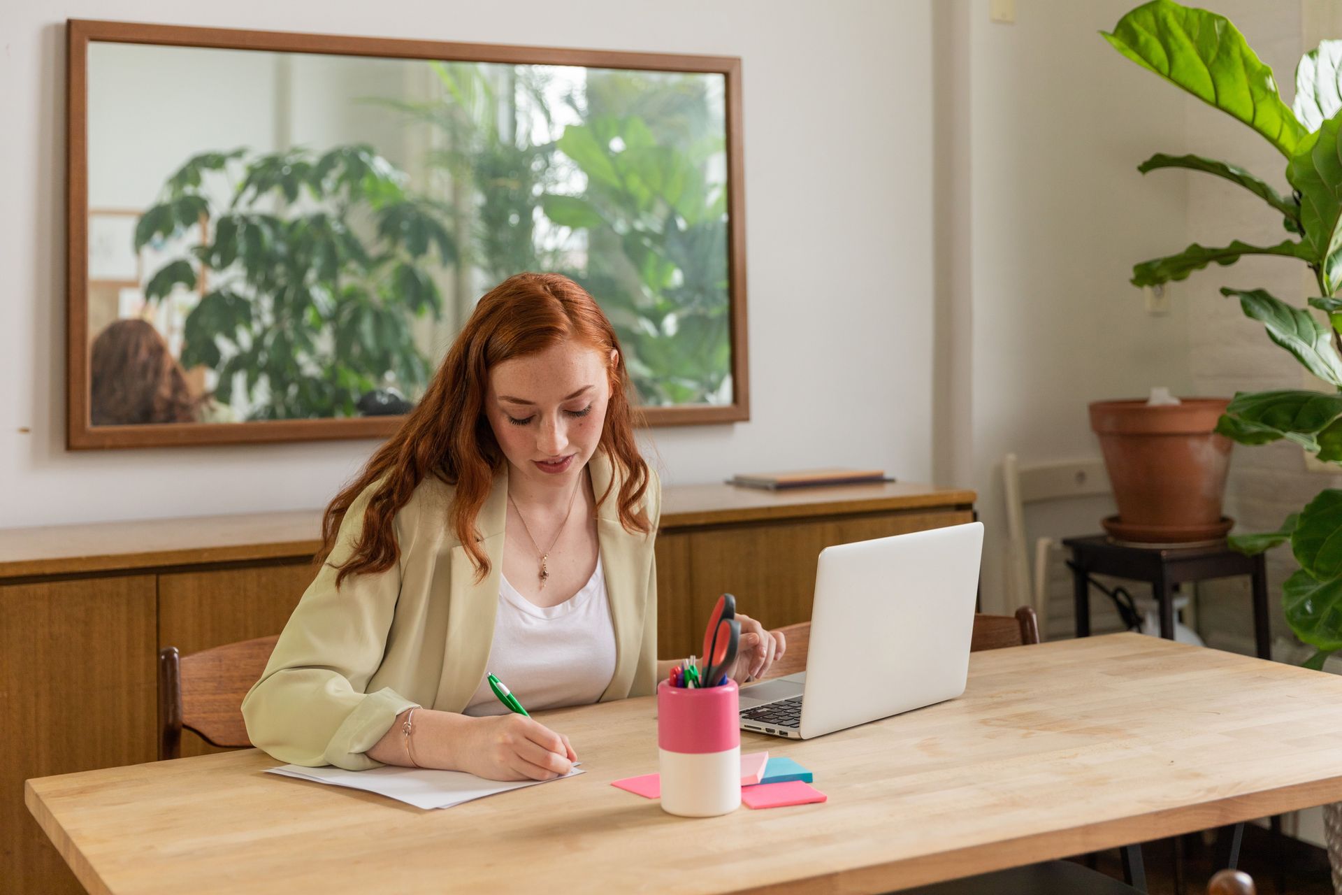 Woman writing at a wooden table with a laptop, pen holder, and notebook in a bright room with plants and a mirror.
