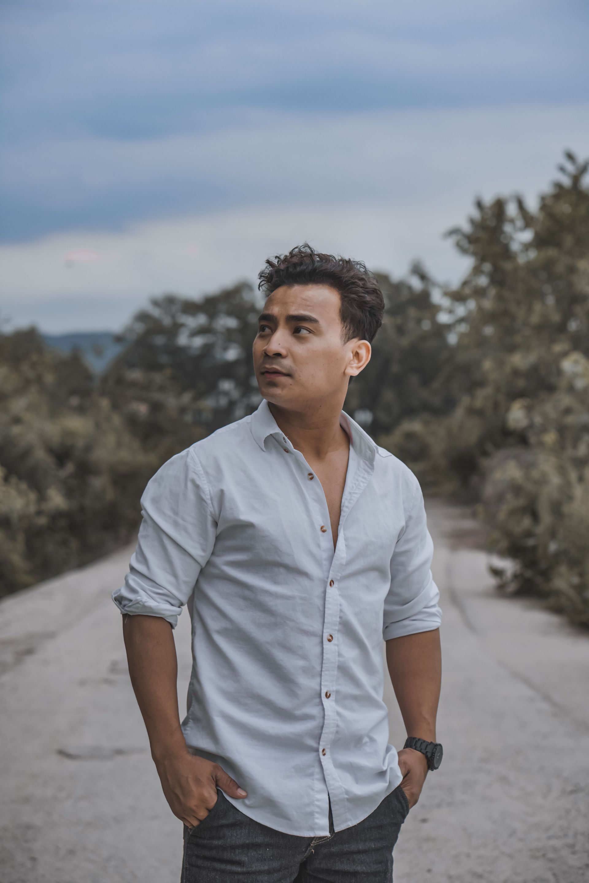 Man in a light blue shirt standing on a road, looking off to the side with trees and cloudy sky behind him