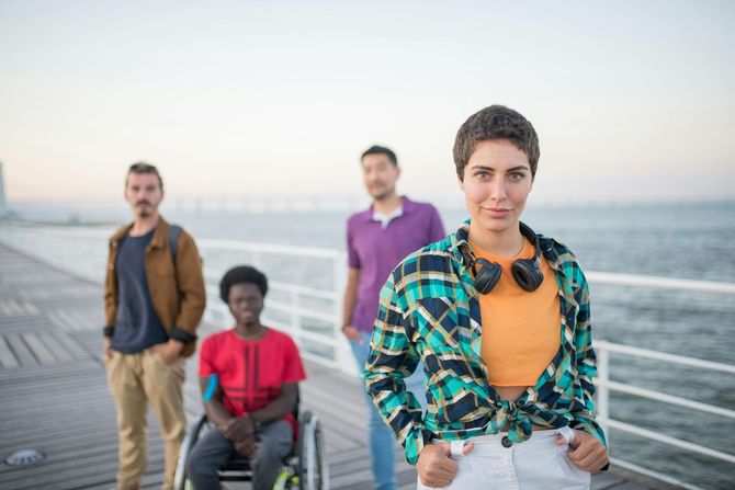 Four people on a waterfront boardwalk, with a woman in a plaid shirt in the foreground and city skyline behind.