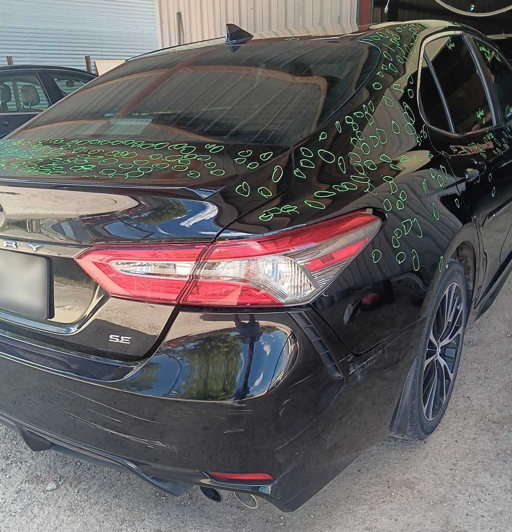 Black Toyota Camry with green markings, likely hail damage, parked outdoors.