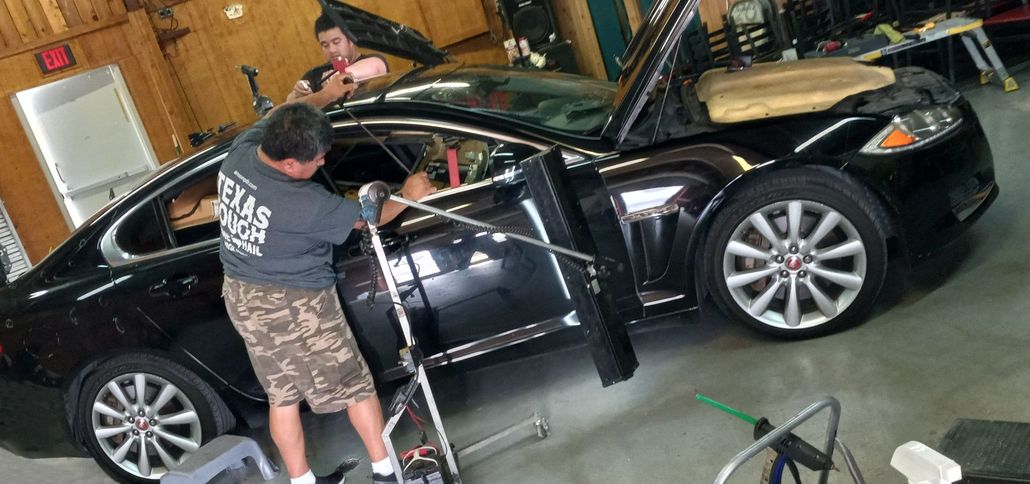 Two people working on a black car in a garage. One inside the car, one outside, both by open doors.