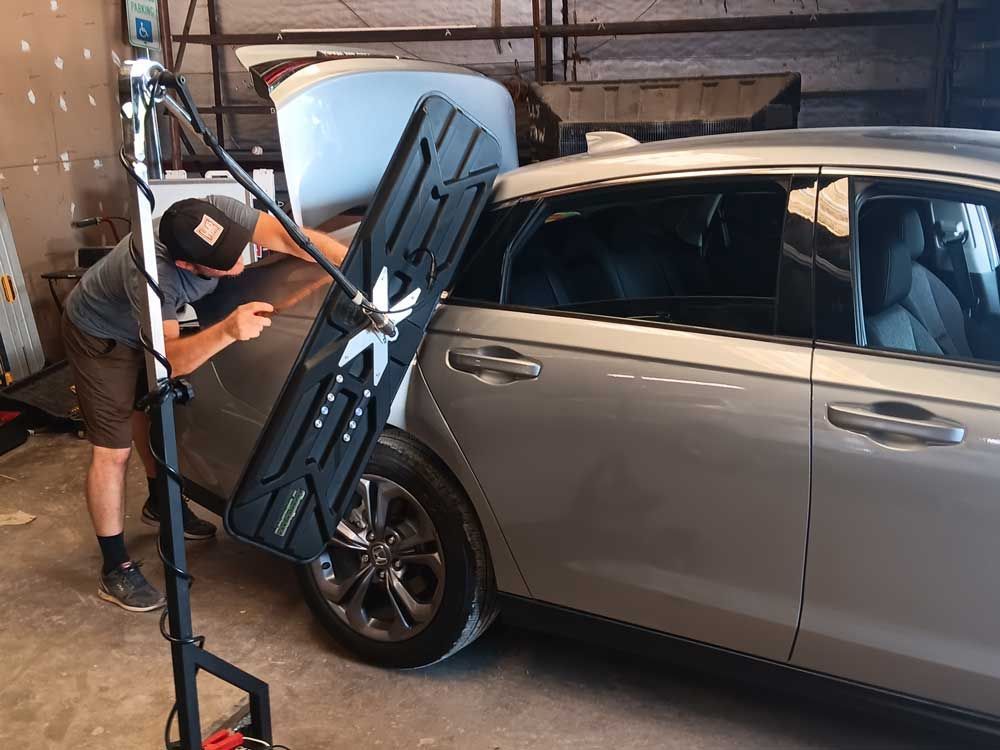 Person using a dent repair light to inspect car damage on a gray sedan's side panel in a garage.