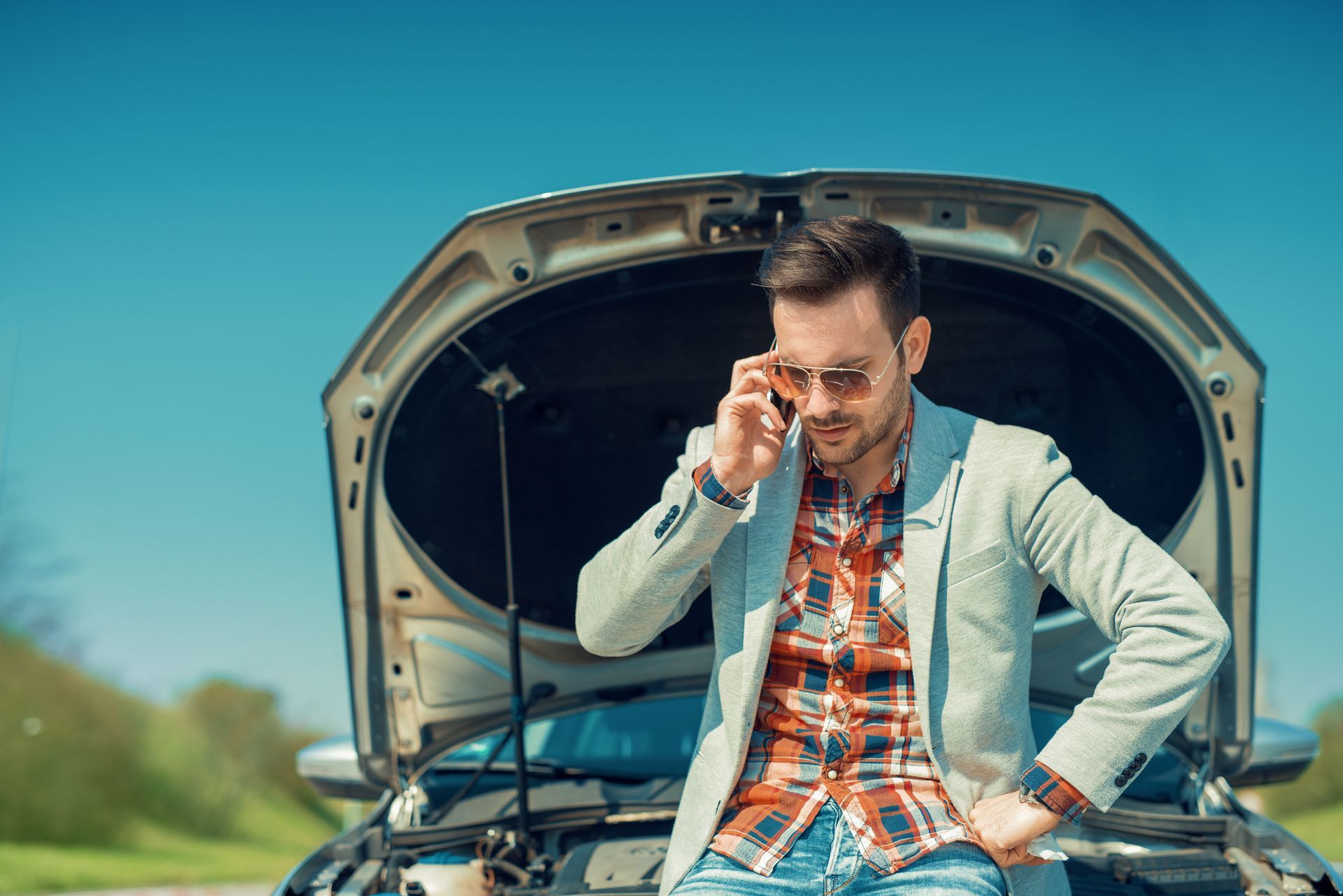 A man stands by his broken car, speaking on his cell phone, seeking 24 Hour Auto Jumpstarts