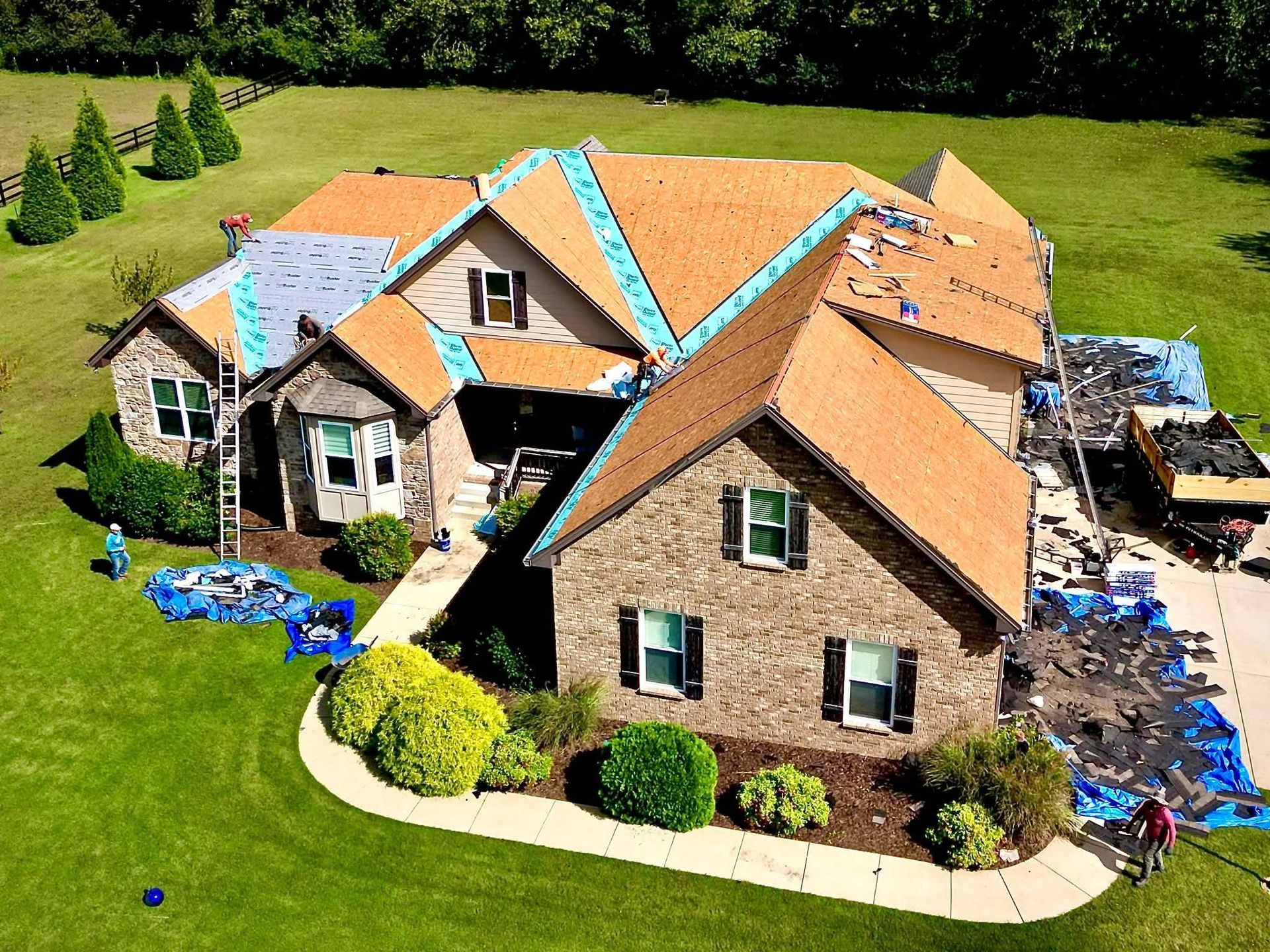 House roof being replaced; workers on roof; brown shingles, blue tarp, green yard.