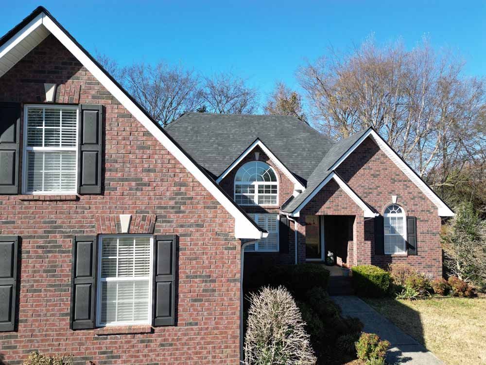 Red brick house with black shutters and a dark gray roof against a blue sky.