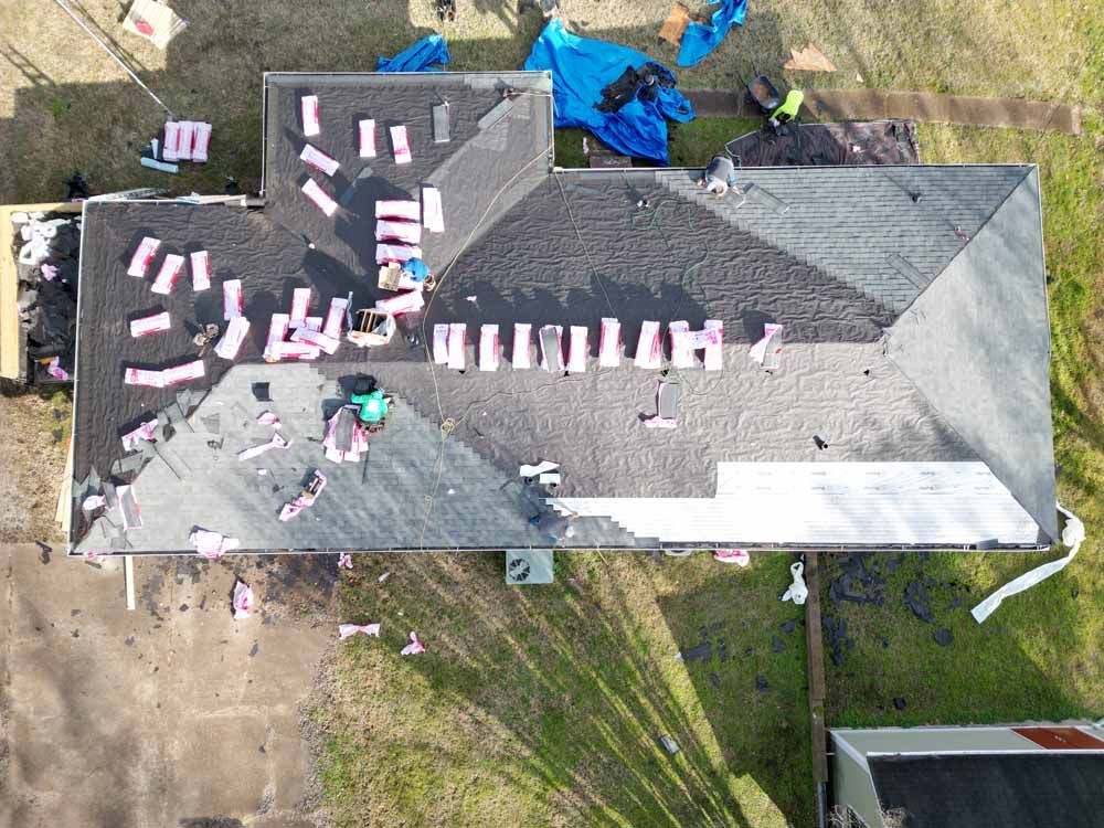 Overhead view of roof with multiple workers. Shingles are being replaced. Pink insulation debris scattered around.