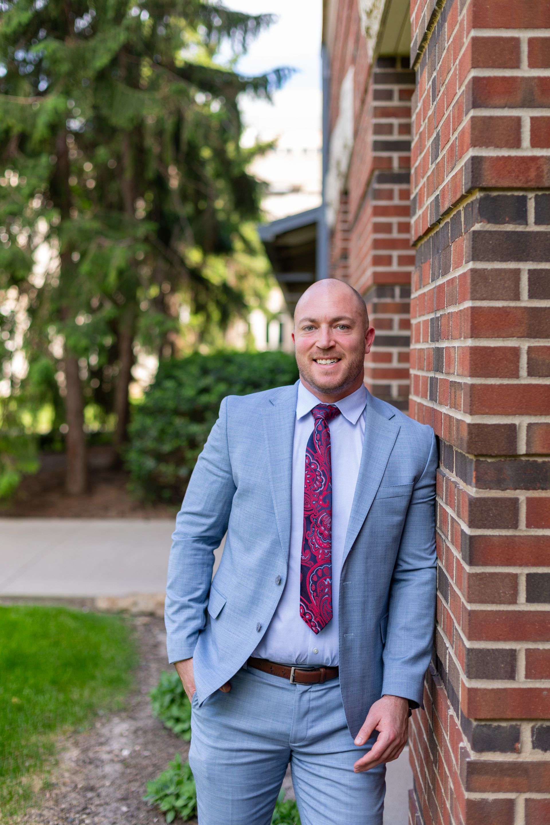 Man in a light blue suit leaning against a brick wall outdoors, smiling.