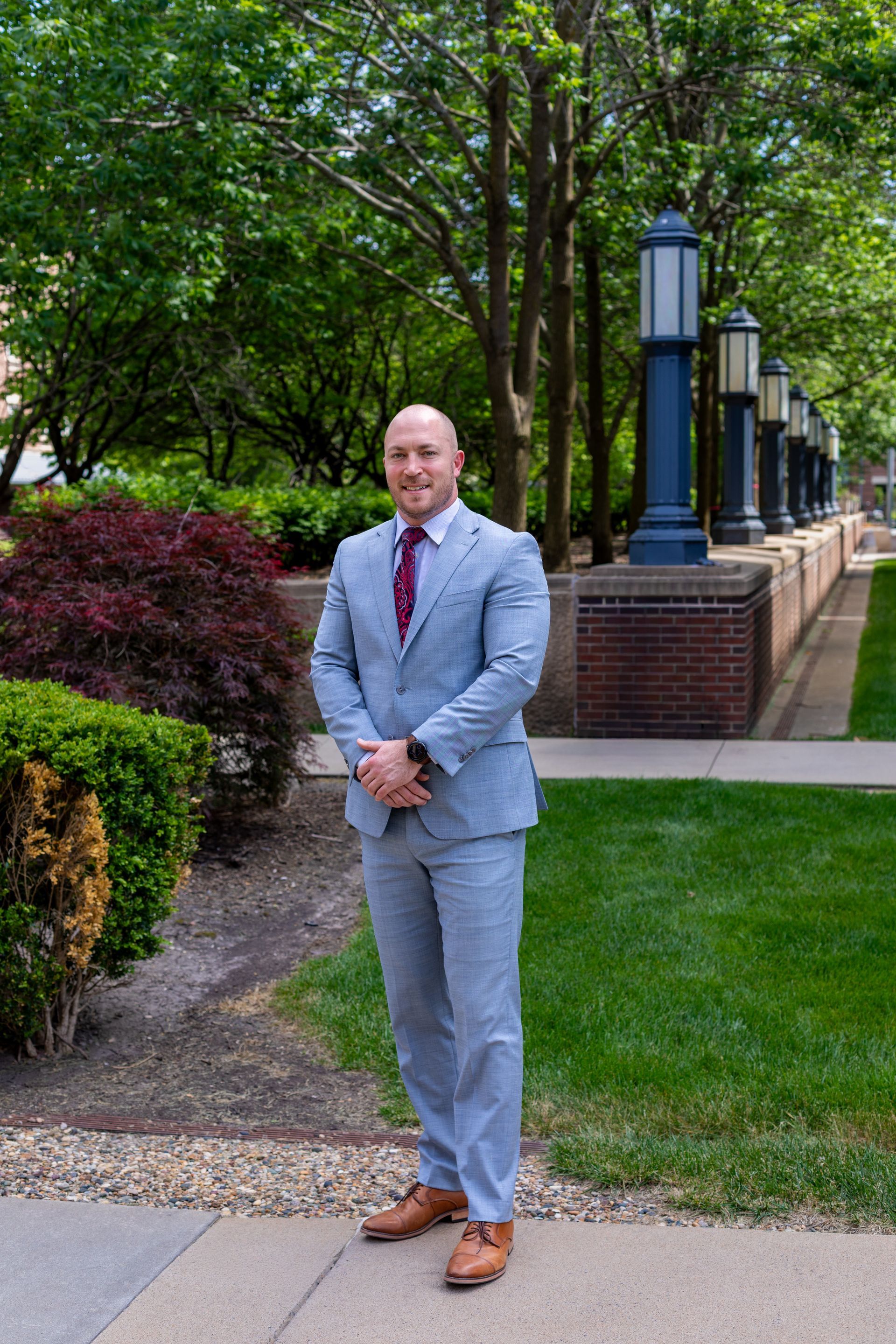 Man in a light gray suit standing on a landscaped walkway beside trees and brick columns.