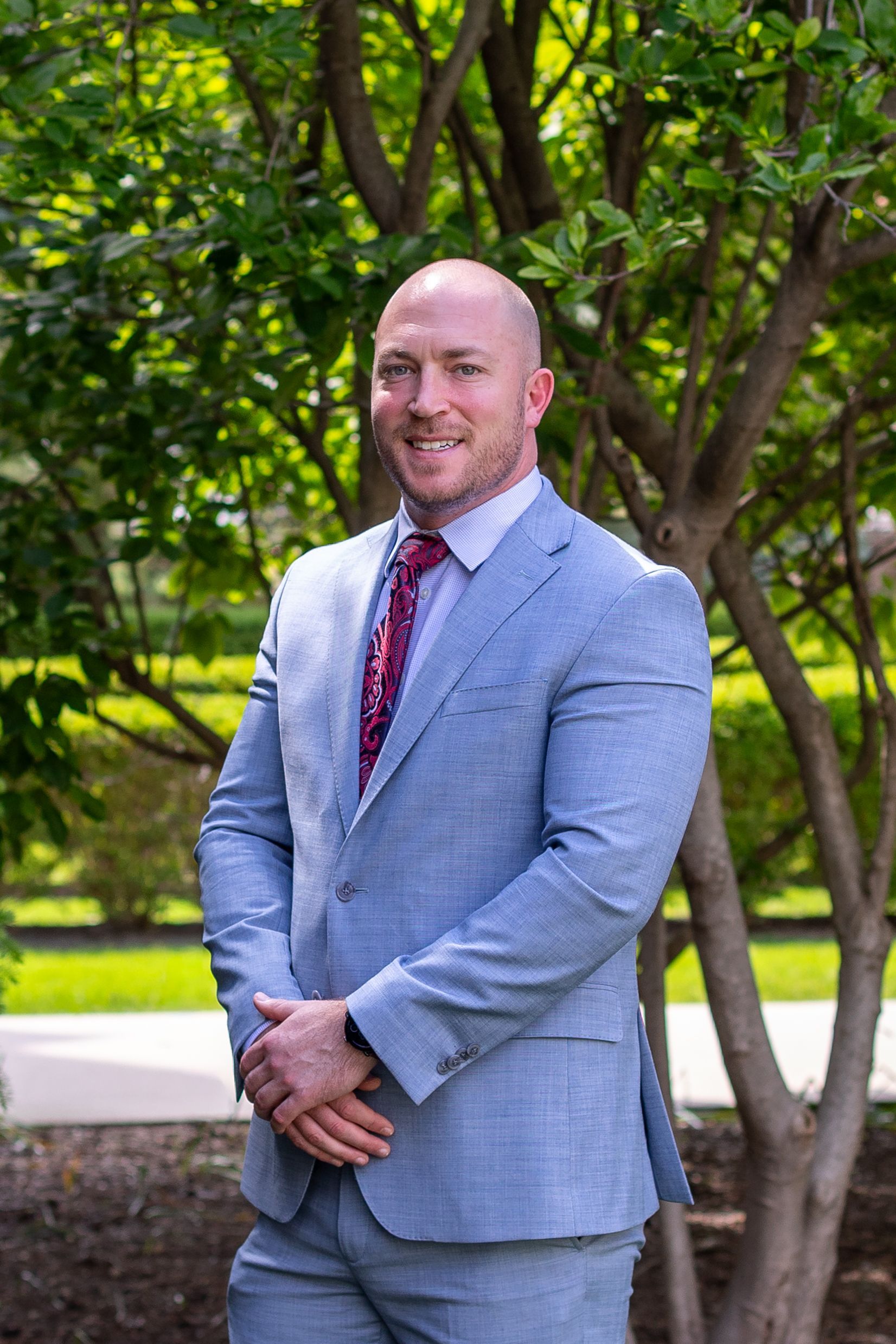 Man in a light blue suit and red tie standing outdoors in a green park, smiling at the camera