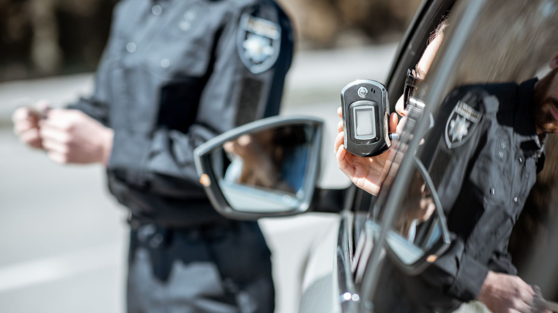 Police officer checking a driver beside a black car, close-up of a hand-held device near the side mirror