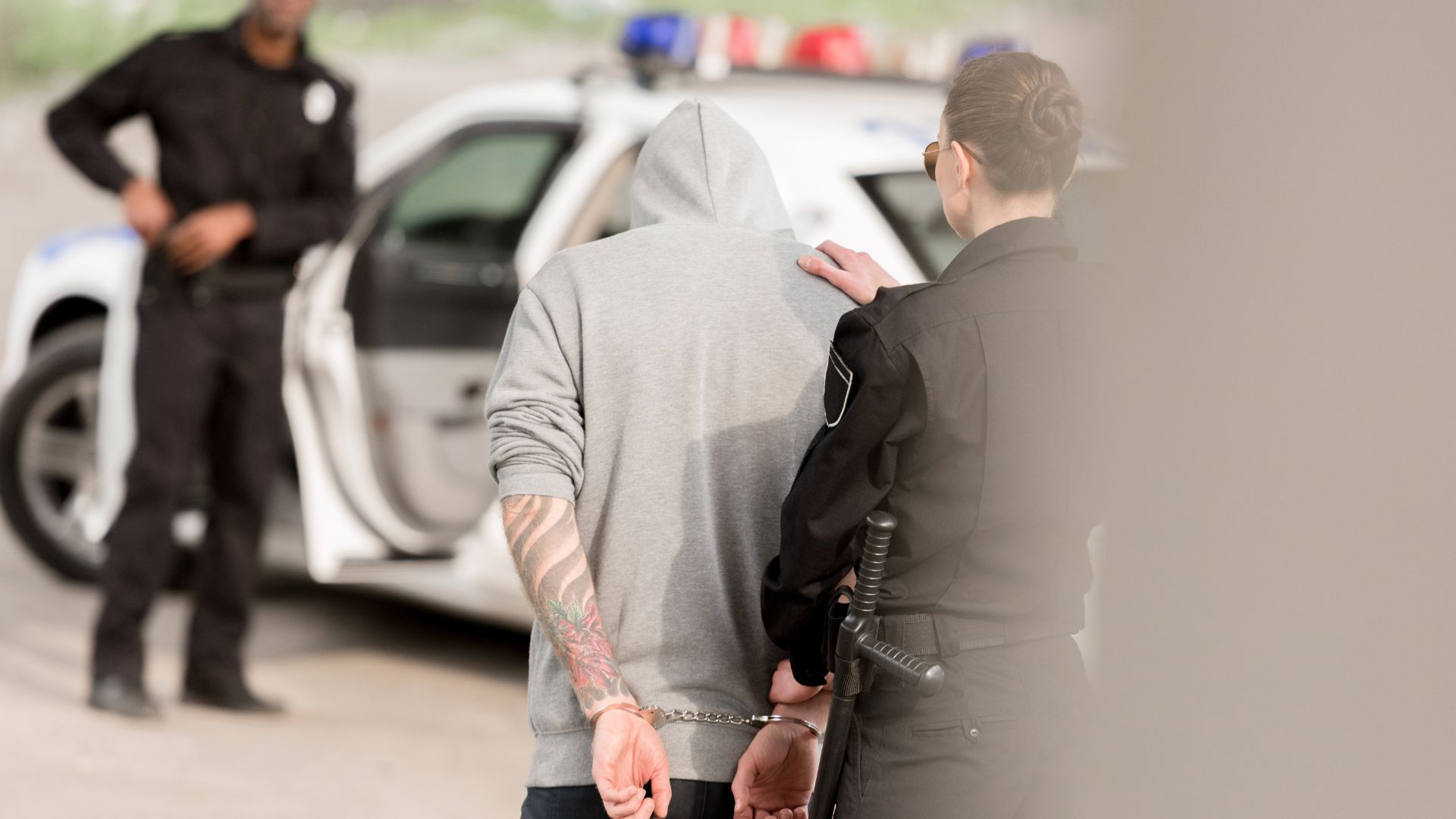 Handcuffed suspect in gray hoodie escorted by police beside a white car