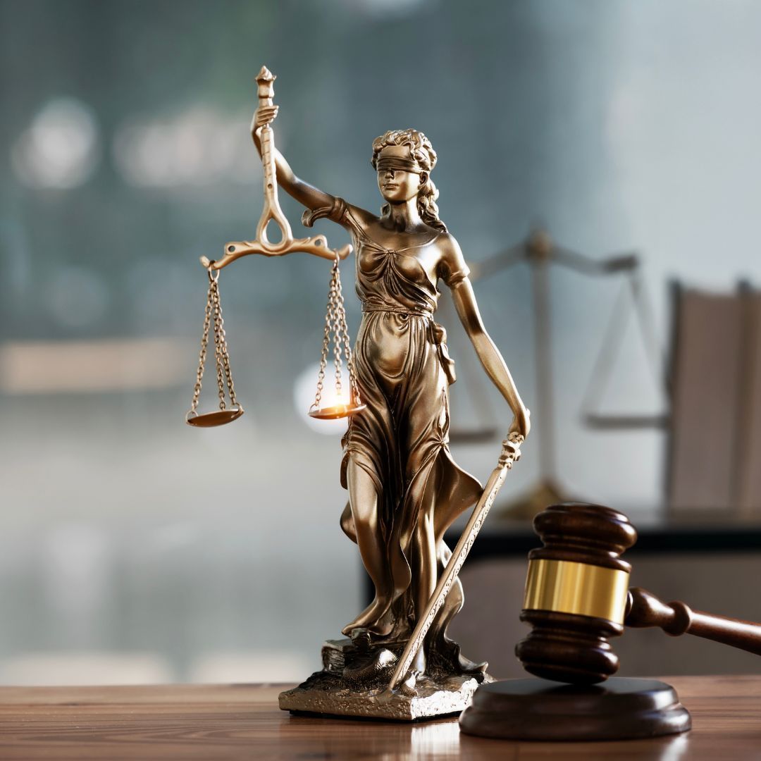Justice statue holding scales beside a judge’s gavel on a desk, with a blurred courtroom background