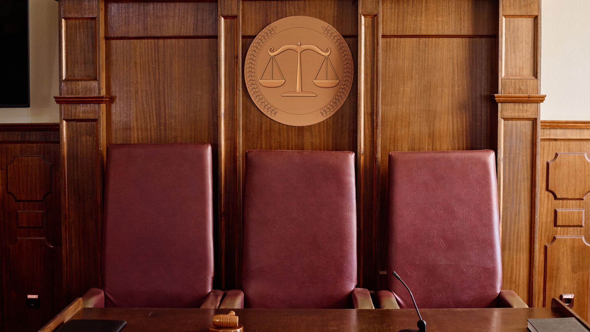 Wooden courtroom bench with three burgundy chairs and a carved scales-of-justice emblem above them
