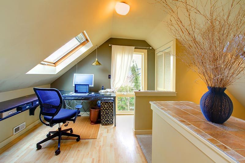 Home office in attic with desk, computer, blue chair, and skylight. Dried plants in blue vase on a counter.
