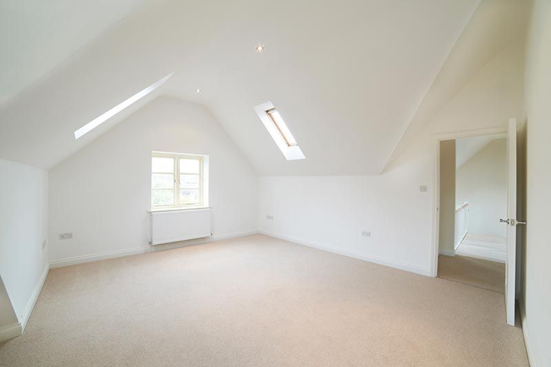 Empty, white-walled room with sloped ceilings and skylights. A small window lets in light, beige carpet.
