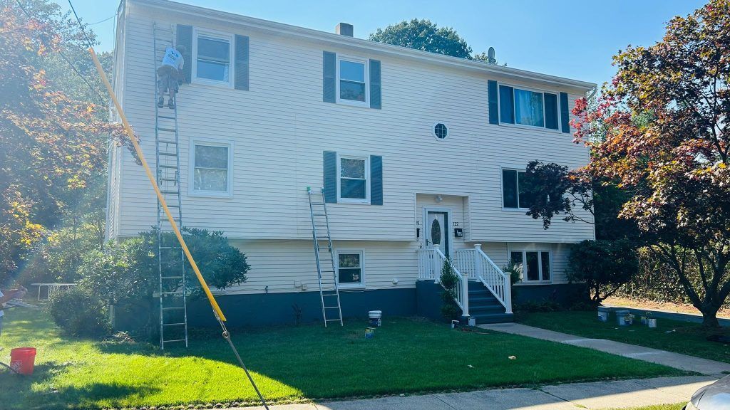 Two-story house being painted; a person on a ladder, using a long pole. Green grass, blue sky.