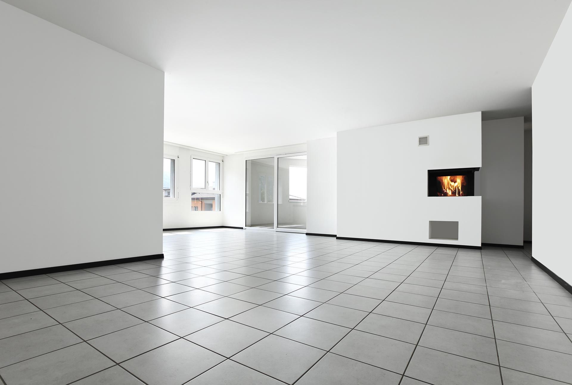 Empty, bright, modern living room with fireplace. White walls, gray tiled floor, and natural light from windows.