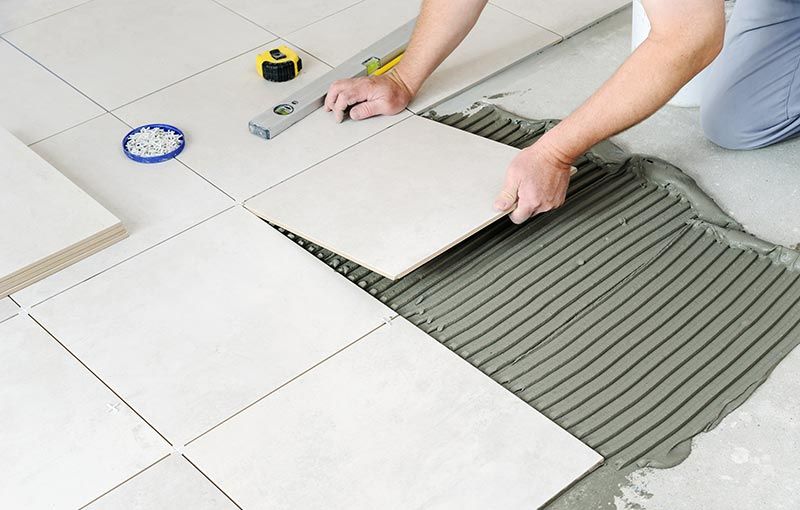 Person laying white tiles on a floor, using a level and trowel to apply adhesive.